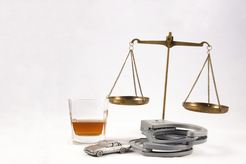 The scales of justice behind a glass of whiskey, a pair of handcuffs and a metal car