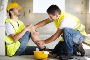A construction worker is holding his leg in pain and another construction workers is aiding him.