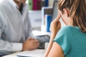 A woman is speaking to a doctor while holding her head in pain.